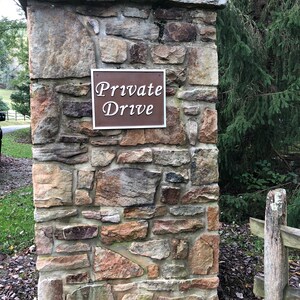 以下が含まれることがあります： 「Private Drive」と書かれた茶色の看板がついた石の柱。看板は柱にネジで固定されています。柱は荒削りの石でできており、緑の葉で囲まれています。