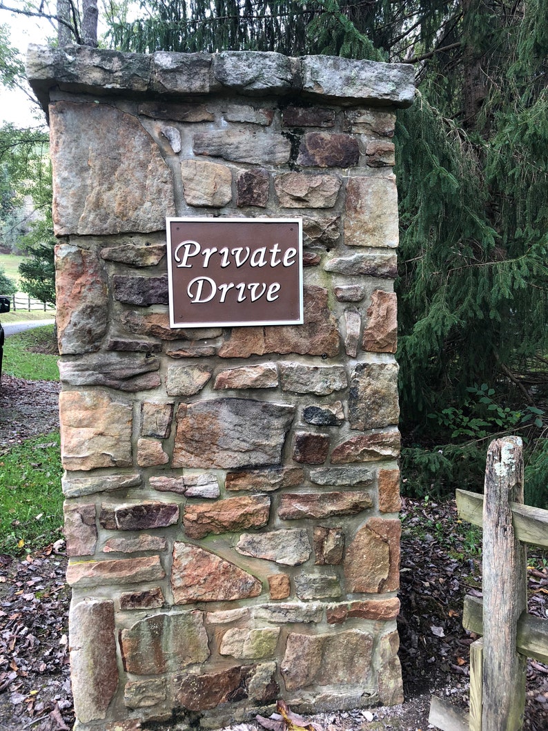 以下が含まれることがあります： 「Private Drive」と書かれた茶色の看板がついた石の柱。看板は柱にネジで固定されています。柱は荒削りの石でできており、緑の葉で囲まれています。