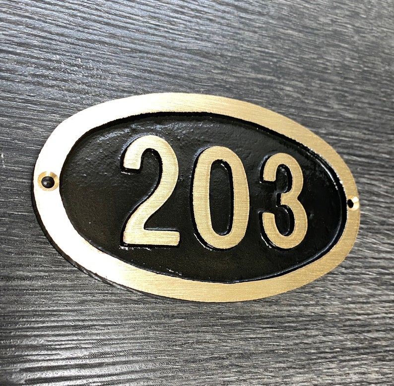May include: A gold oval-shaped house number plaque with black lettering. The number 203 is displayed on the plaque.