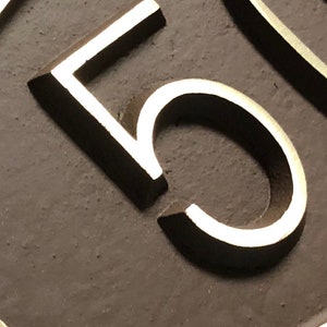 May include: Close-up of a silver number "5" on a dark brown oval background.