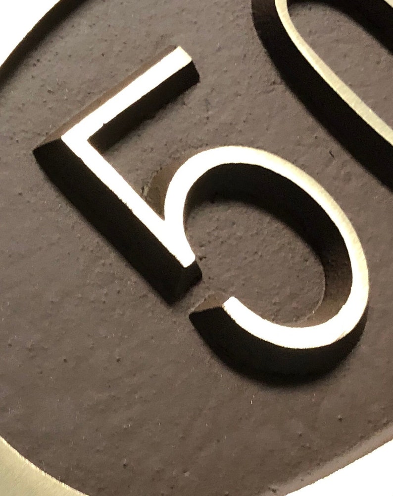 May include: Close-up of a silver number "5" on a dark brown oval background.