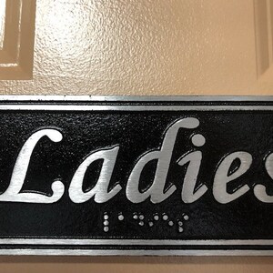 May include: A black and silver sign with the word "Ladies" in silver lettering. The sign has a raised border and braille lettering.