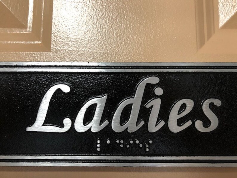 May include: A black and silver sign with the word "Ladies" in silver lettering. The sign has a raised border and braille lettering.