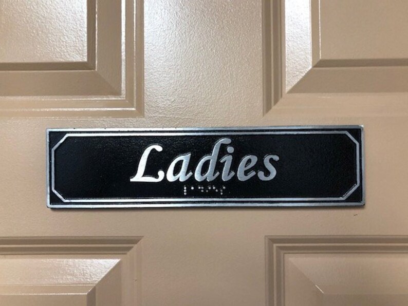 May include: A black and silver sign with the word "Ladies" in silver lettering on a beige door.