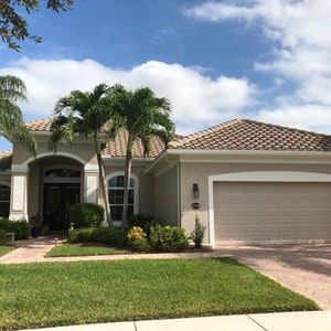 May include: A beige single-storey house with a brown tiled roof, a white garage door, and a pathway leading to the front door. The house is surrounded by lush green grass and palm trees.