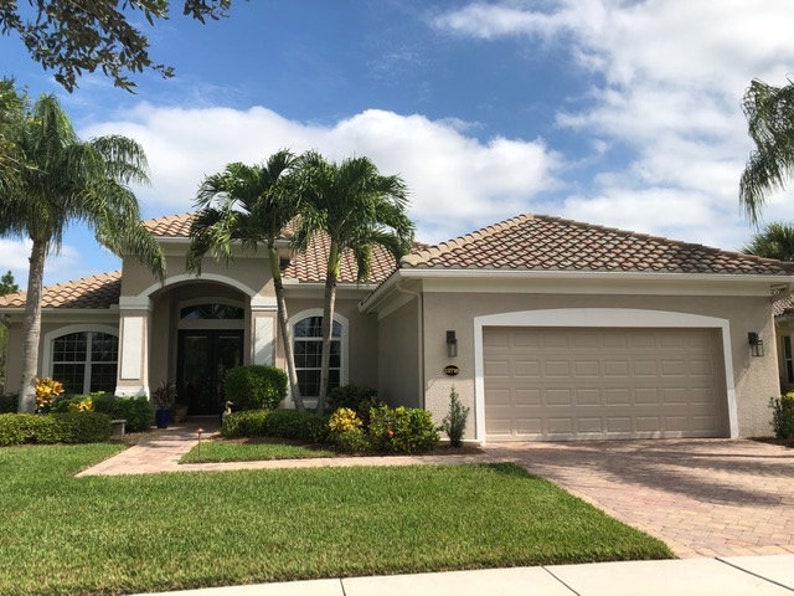 May include: A beige single-storey house with a brown tiled roof, a white garage door, and a pathway leading to the front door. The house is surrounded by lush green grass and palm trees.