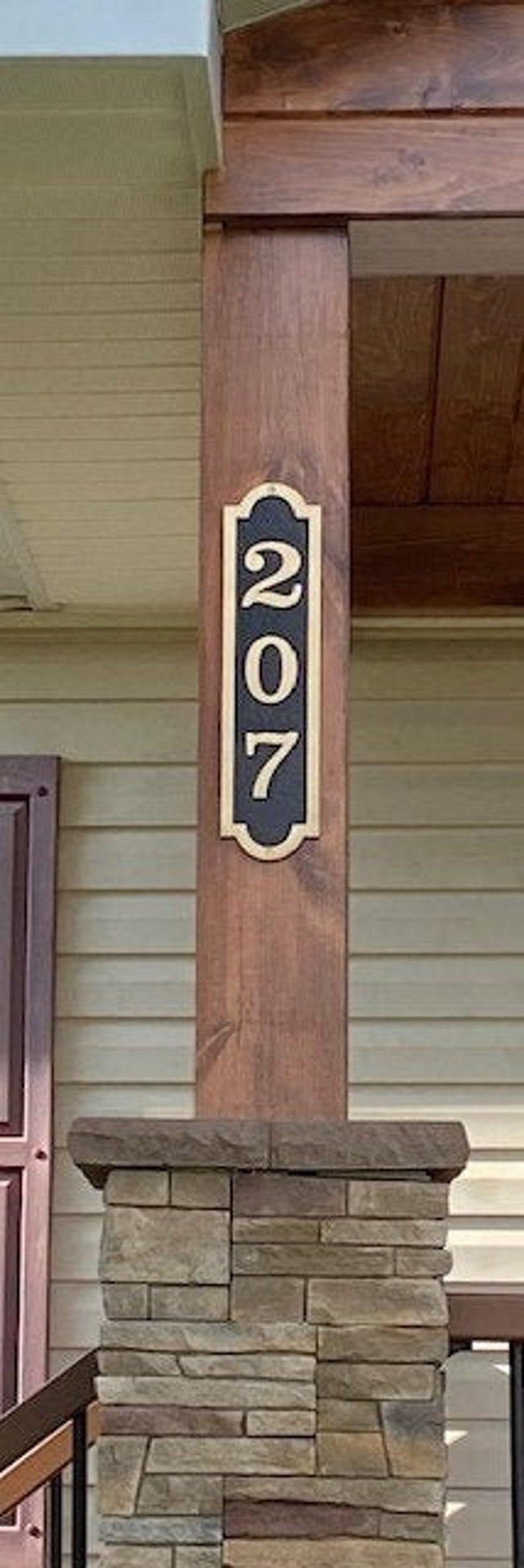 May include: A black and gold house number sign with the number 207 on a wooden post. The post is attached to a stone pillar.