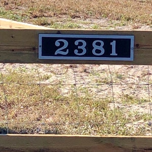 May include: A rectangular address plaque with the number "2381" in white against a black background. The plaque is mounted on a wooden fence, with a wire mesh fence below. The background features dry grass and a sunny outdoor setting.