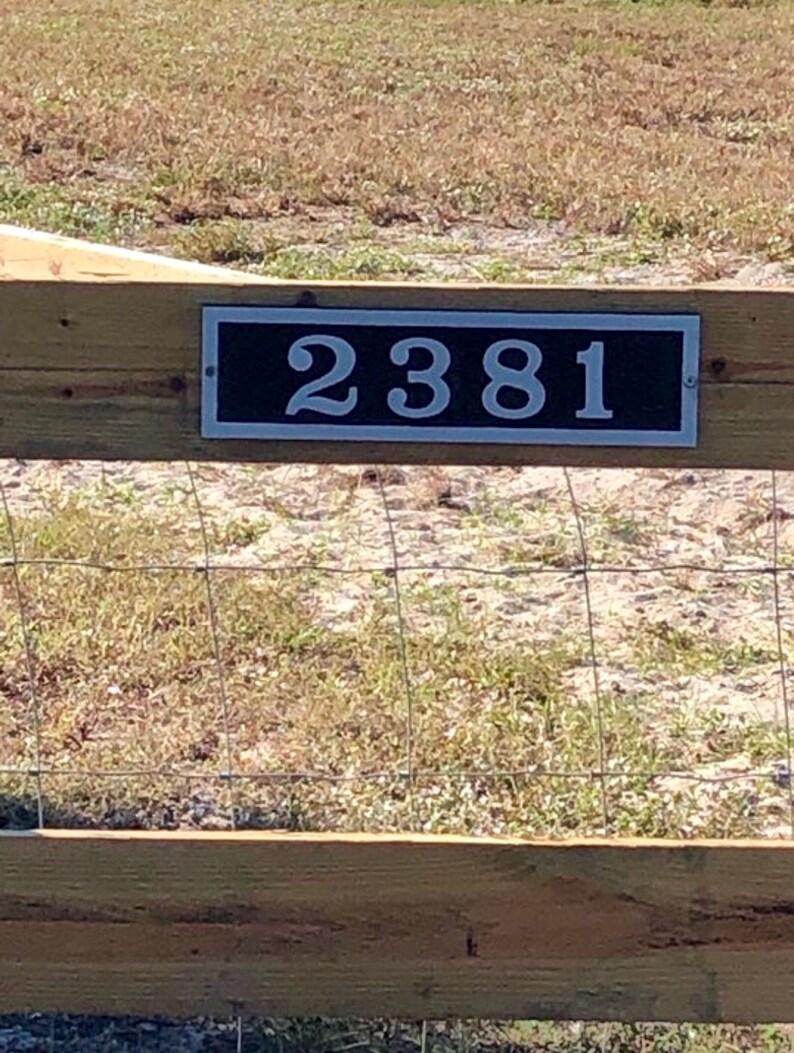 May include: A rectangular address plaque with the number "2381" in white against a black background. The plaque is mounted on a wooden fence, with a wire mesh fence below. The background features dry grass and a sunny outdoor setting.