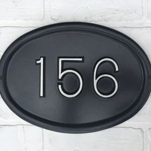 May include: Black oval-shaped house number sign with white numbers "156" on a white brick wall background.