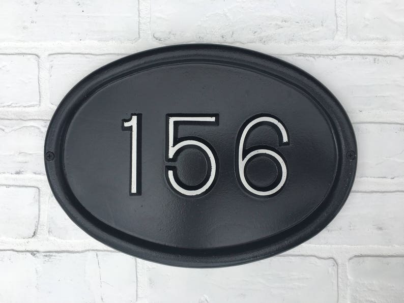 May include: Black oval-shaped house number sign with white numbers "156" on a white brick wall background.