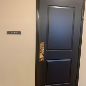 May include: A black door with a gold handle and a sign that says "Ladies" on a beige wall.