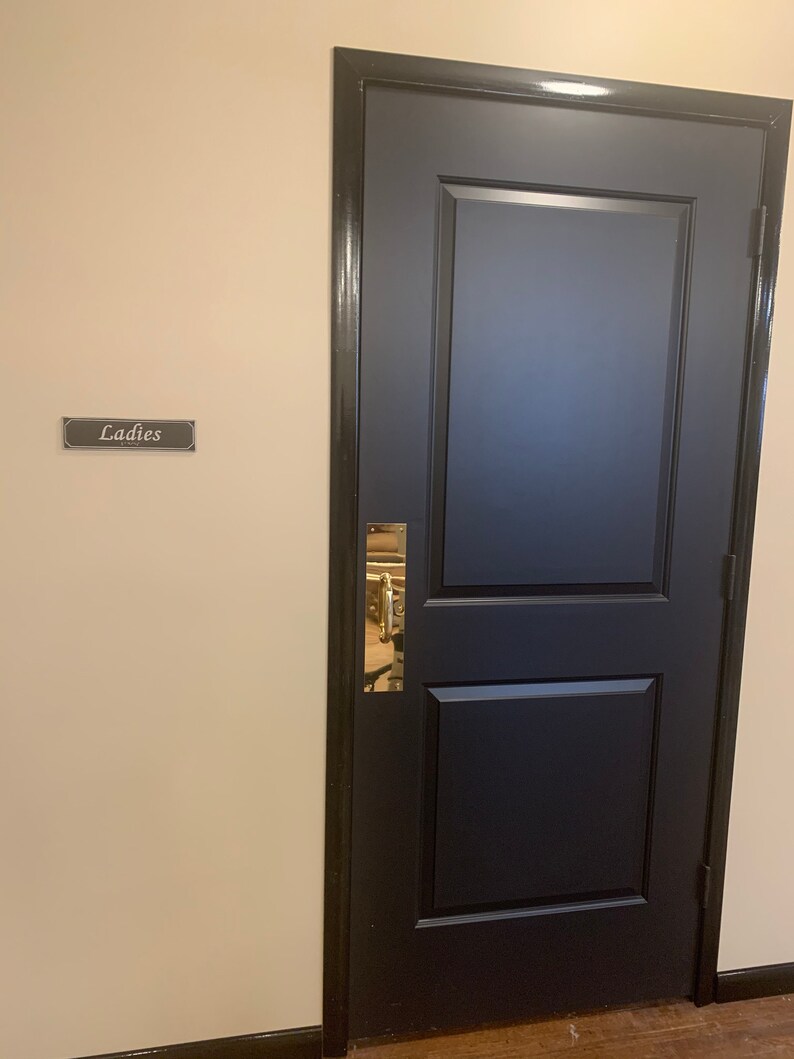 May include: A black door with a gold handle and a sign that says "Ladies" on a beige wall.