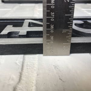 以下が含まれることがあります： 黒と銀の金属製の家の番号プレートのクローズアップ。プレートには「04」という数字が刻印されています。定規で測定したところ、幅が9.9cmであることがわかります。