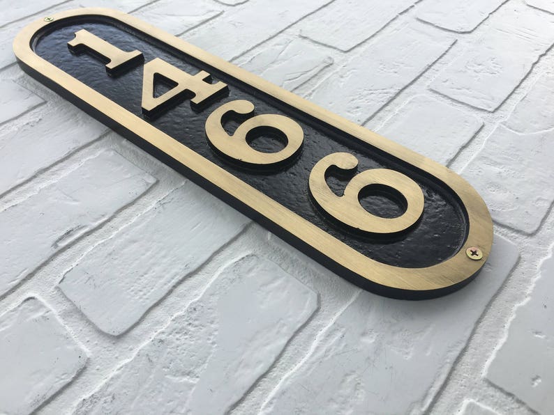 May include: A black and gold oval-shaped house number sign with the number 146 on it. The sign is mounted on a white brick wall.