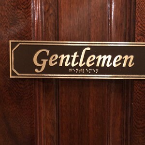 May include: A brown rectangular sign with a gold border and the word "Gentlemen" in gold lettering. The sign also includes braille lettering.