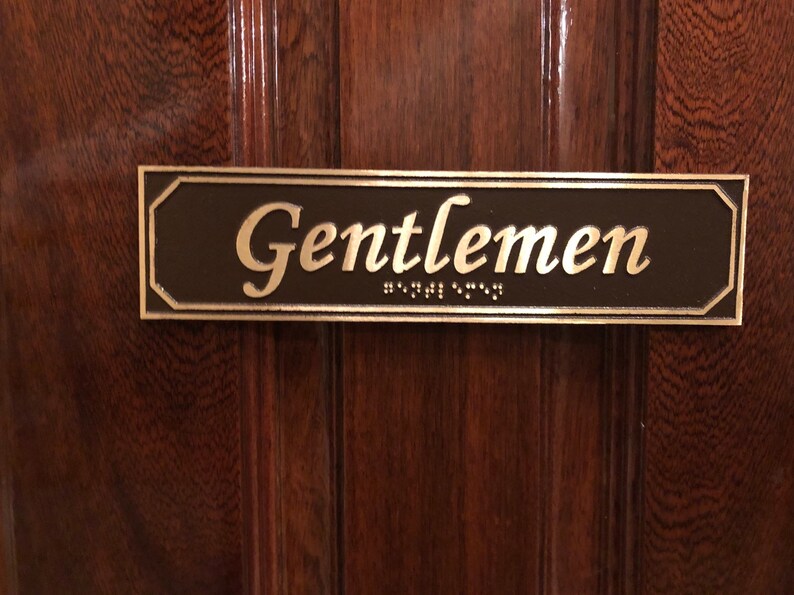May include: A brown rectangular sign with a gold border and the word "Gentlemen" in gold lettering. The sign also includes braille lettering.
