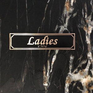 May include: A black sign with gold trim that reads "Ladies" in gold lettering. The sign also has braille lettering.
