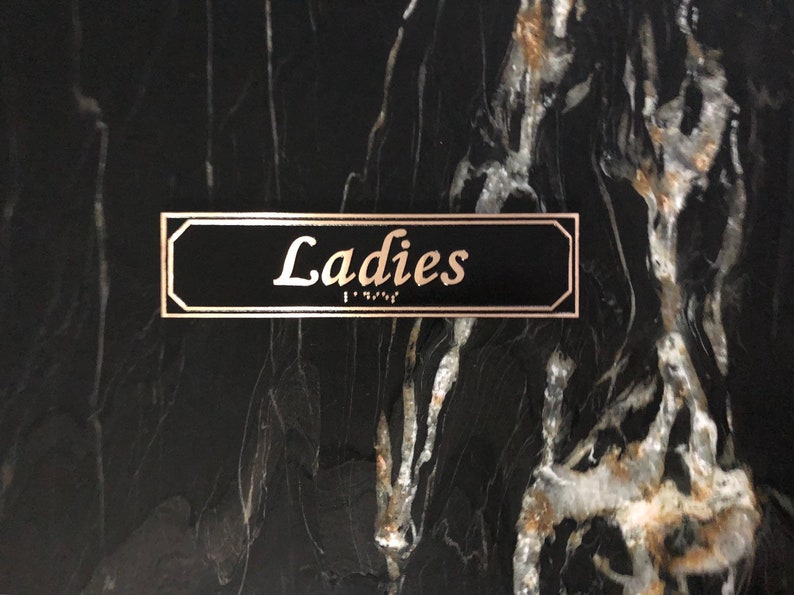 May include: A black sign with gold trim that reads "Ladies" in gold lettering. The sign also has braille lettering.