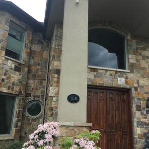 May include: A stone house with a wooden door and a black oval sign with the number 156 on it. There are pink flowers in front of the house.