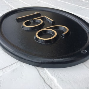 May include: Black oval house number plaque with gold-colored numbers "156" on a white brick wall.