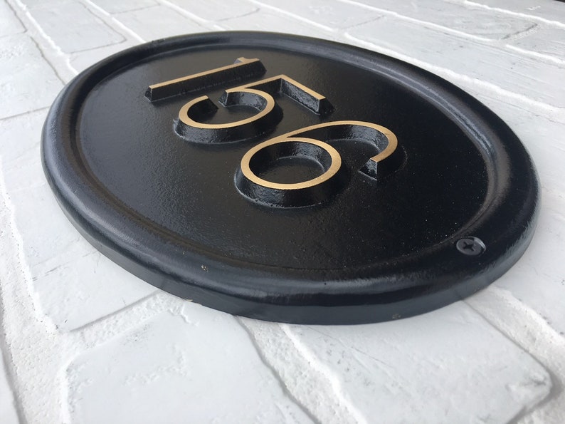 May include: Black oval house number plaque with gold-colored numbers "156" on a white brick wall.