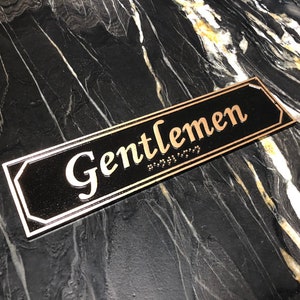 May include: A black rectangular sign with gold trim and the word "Gentlemen" in gold lettering. The sign also has braille lettering.