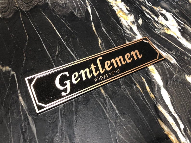 May include: A black rectangular sign with gold trim and the word "Gentlemen" in gold lettering. The sign also has braille lettering.