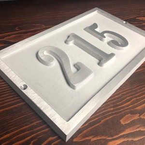 May include: White rectangular house number sign with a silver frame and the number 215 embossed in silver.