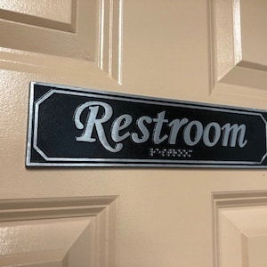 May include: A black rectangular sign with silver lettering that reads "Restroom" with braille below the text.