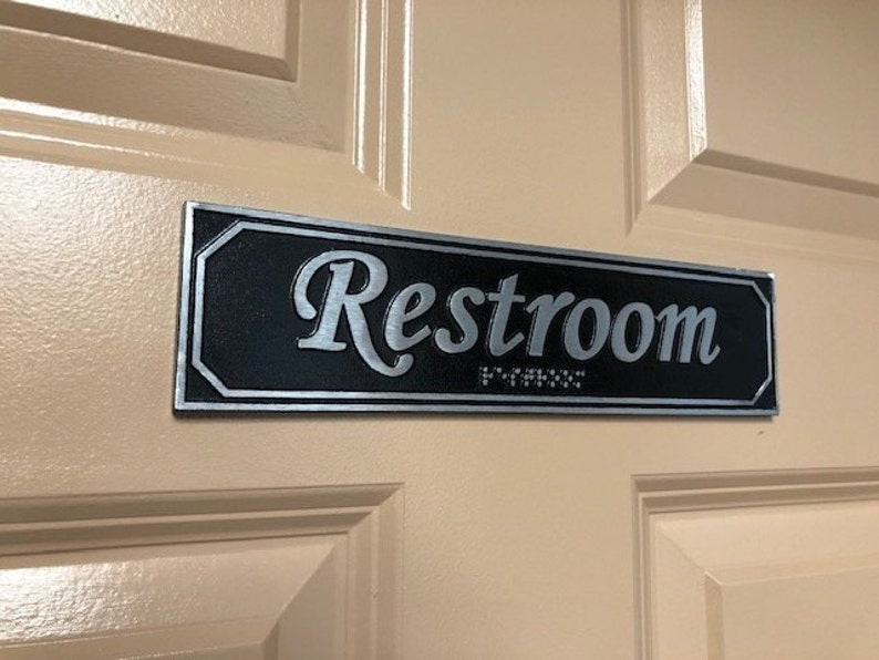 May include: A black rectangular sign with silver lettering that reads "Restroom" with braille below the text.
