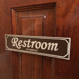 May include: A brown rectangular sign with a white border and the word "Restroom" in white cursive lettering. The sign is mounted on a wooden door.