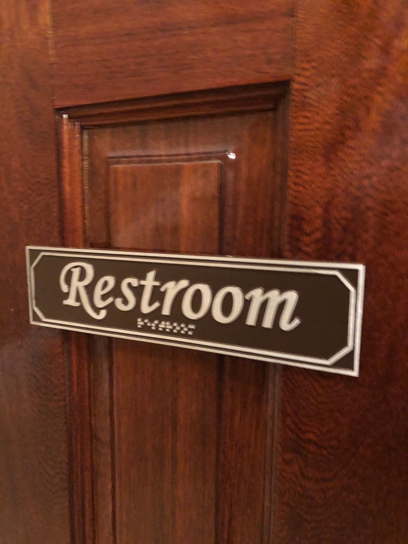 May include: A brown rectangular sign with a white border and the word "Restroom" in white cursive lettering. The sign is mounted on a wooden door.