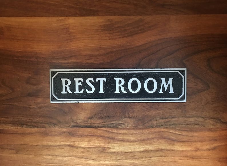 Pu&ograve; includere: Un cartello nero e argento con la scritta "REST ROOM" in lettere bianche.