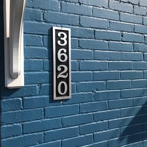 Puede incluir: Un letrero de n&uacute;mero de casa de metal plateado con el n&uacute;mero 3620 en una pared de ladrillo azul.