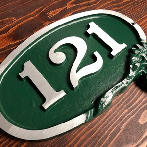 May include: Oval-shaped house number sign with the number 121 in silver lettering on a green background. The sign has a silver border and an acorn design on the right side.