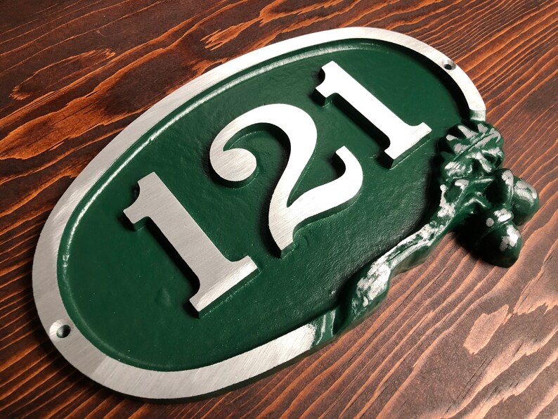 May include: Oval-shaped house number sign with the number 121 in silver lettering on a green background. The sign has a silver border and an acorn design on the right side.