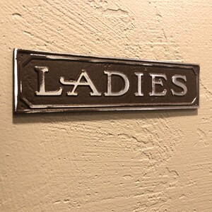 K&ouml;nnte beinhalten: Ein braunes Metallschild mit silbernen Buchstaben, auf dem "LADIES" steht.