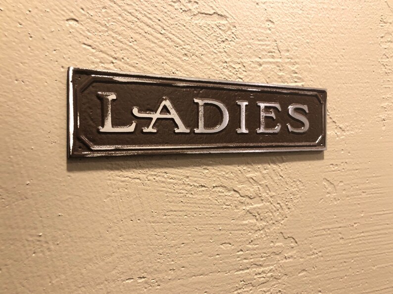 K&ouml;nnte beinhalten: Ein braunes Metallschild mit silbernen Buchstaben, auf dem "LADIES" steht.