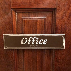 May include: A brown wooden door with a silver plaque that reads "Office" in white lettering. The plaque also has braille lettering.