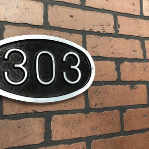 May include: A silver oval-shaped house number sign with a black background and the number 303 in white.