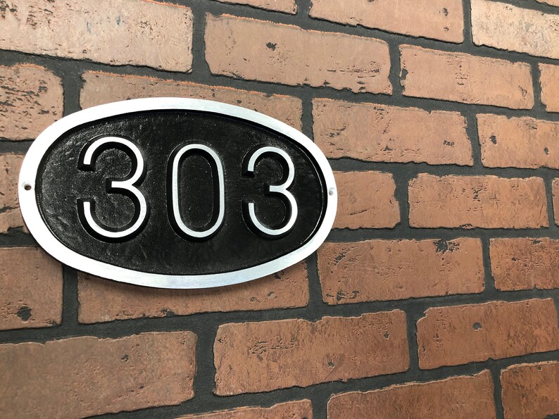 May include: A silver oval-shaped house number sign with a black background and the number 303 in white.