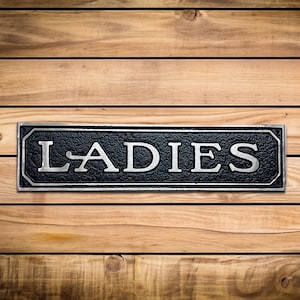 K&ouml;nnte beinhalten: Schwarzes und silbernes Metallschild mit dem Wort "LADIES" in wei&szlig;en Buchstaben.