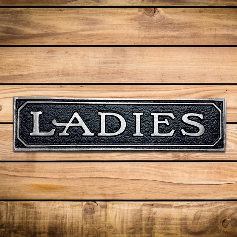 K&ouml;nnte beinhalten: Schwarzes und silbernes Metallschild mit dem Wort "LADIES" in wei&szlig;en Buchstaben.