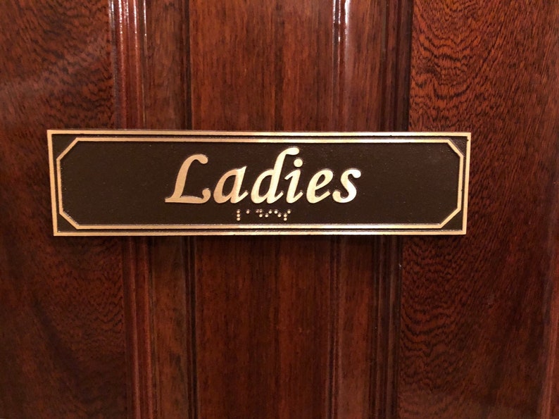 May include: A brown rectangular sign with a gold border and the word "Ladies" in gold lettering. The sign also has braille lettering.