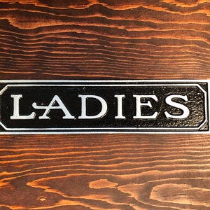 May include: A black and silver sign with the word "LADIES" in white lettering.