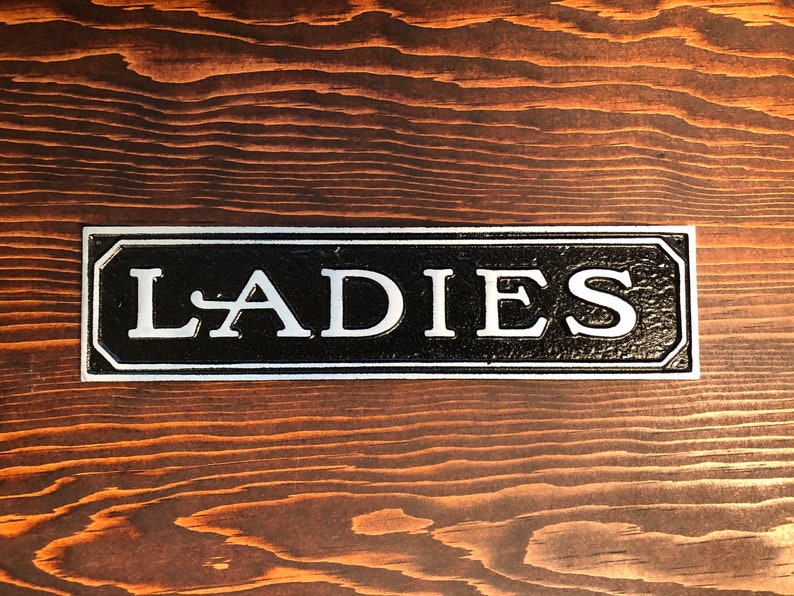 May include: A black and silver sign with the word "LADIES" in white lettering.