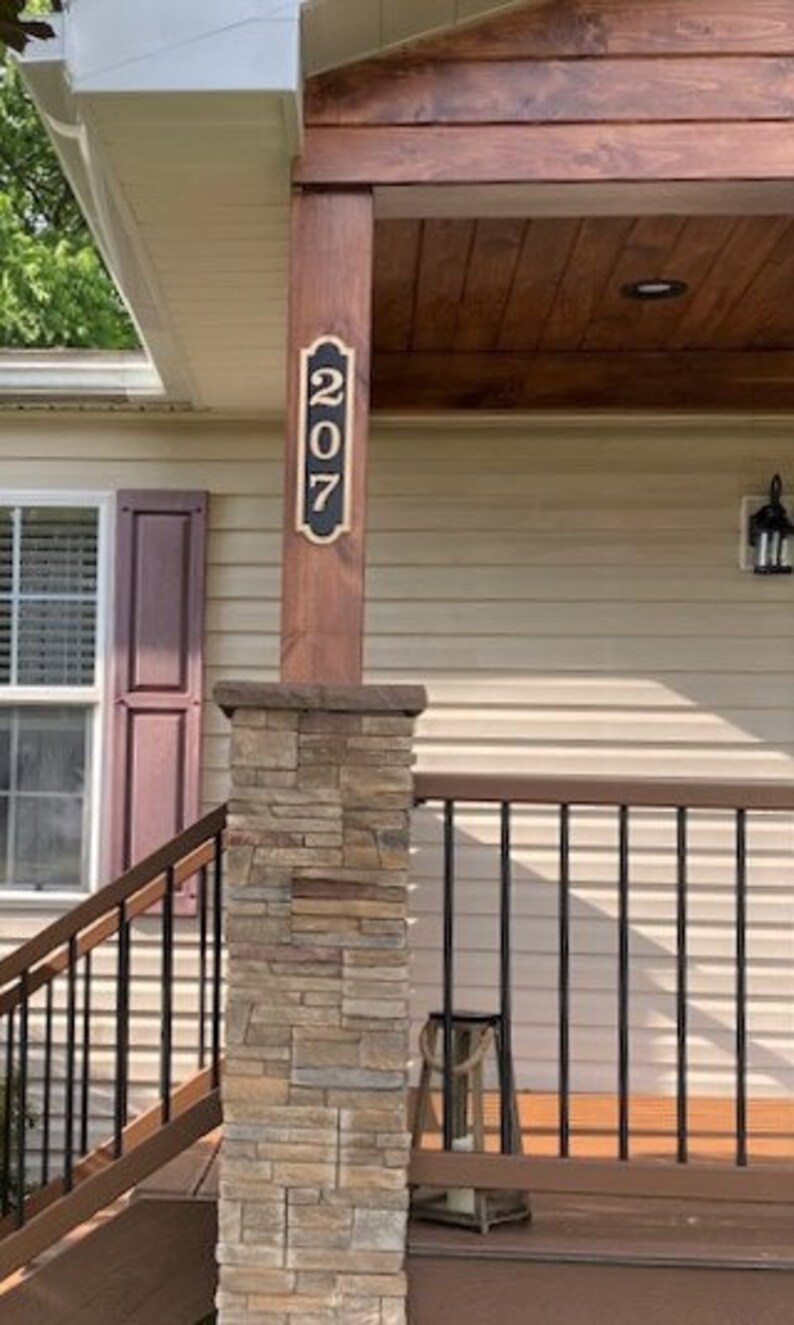May include: A house number sign with the number 207 on it. The sign is mounted on a wooden post and has a black background with gold trim. The post is made of stone and is surrounded by a wooden railing.