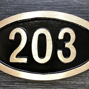 May include: Oval-shaped brass house number plaque with a black background and gold-colored numerals. The number 203 is displayed on the plaque.