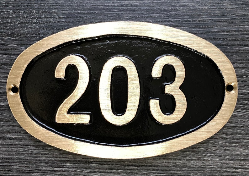 May include: Oval-shaped brass house number plaque with a black background and gold-colored numerals. The number 203 is displayed on the plaque.